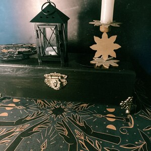 May include: A black wooden box with a gold latch and a black metal lantern with a candle inside. The box is open and the inside is lined with black paper with a gold design of hands reaching up towards a star.