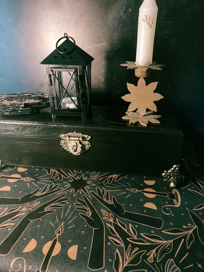 May include: A black wooden box with a gold latch and a black metal lantern with a candle inside. The box is open and the inside is lined with black paper with a gold design of hands reaching up towards a star.