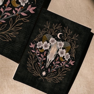 May include: Two black art prints featuring a ram skull surrounded by white flowers, pink leaves, and gold-colored branches. A crescent moon and star are also included in the design. The prints have a textured appearance.