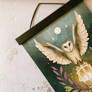 May include: A wall hanging featuring an illustration of a barn owl in flight against a dark green and blue night sky. The owl is depicted with outstretched wings, carrying a glowing lantern. The artwork includes a moon, stars, and floral accents.