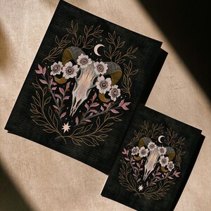 May include: Two black art prints featuring a ram skull with flowers, leaves, and a crescent moon. The artwork has a dark, gothic aesthetic with gold, pink, and white accents. The prints are of different sizes.