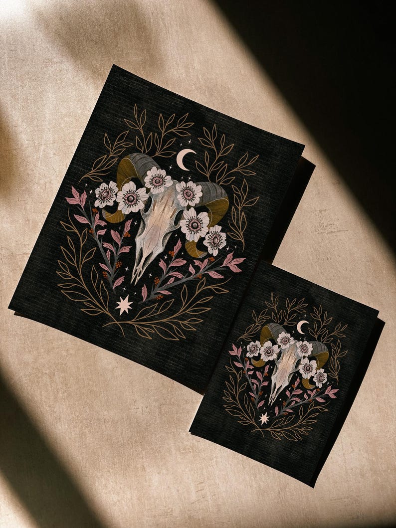 May include: Two black art prints featuring a ram skull with flowers, leaves, and a crescent moon. The artwork has a dark, gothic aesthetic with gold, pink, and white accents. The prints are of different sizes.