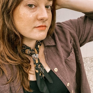 Op de afbeelding: Een vrouw met lang bruin haar draagt een bruin shirt met een zwarte bandana om haar nek. De bandana heeft een goud en zwart patroon. De vrouw draagt een zwarte tanktop onder het shirt. De vrouw heeft een tatoeage op haar pols met de tekst "Lunar".