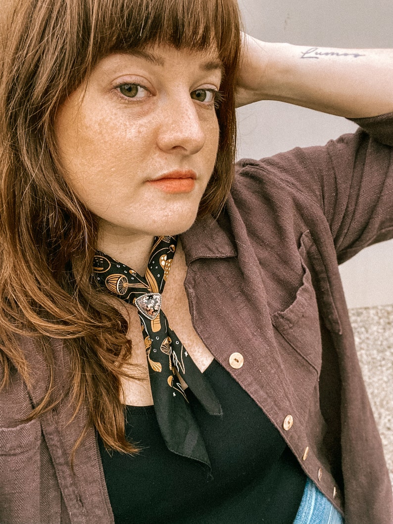 Op de afbeelding: Een vrouw met lang bruin haar draagt een bruin shirt met een zwarte bandana om haar nek. De bandana heeft een goud en zwart patroon. De vrouw draagt een zwarte tanktop onder het shirt. De vrouw heeft een tatoeage op haar pols met de tekst "Lunar".