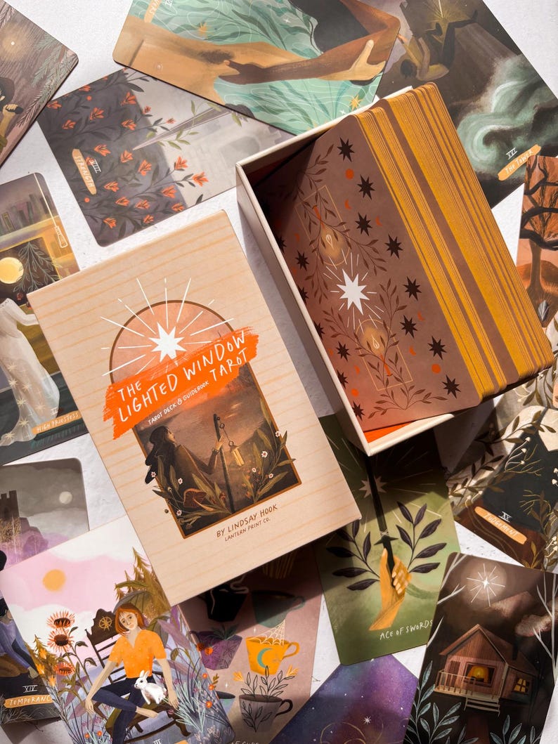 May include: A tarot deck and guidebook set titled "The Lighted Window Tarot" by Lindsay Hook. The wooden box and cards feature illustrations in shades of orange, brown, and green. The cards are scattered around the box, showcasing various artistic designs.