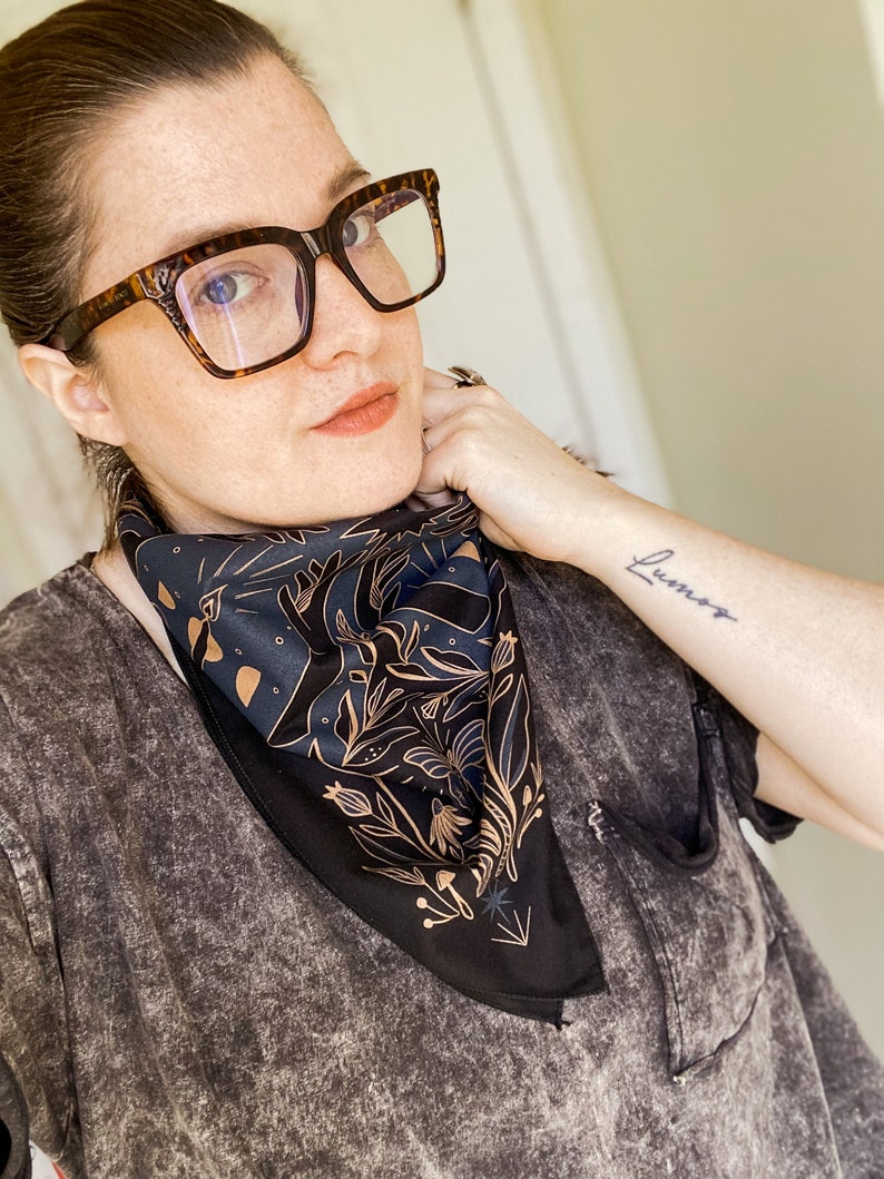 May include: A woman wearing tortoiseshell glasses and a black t-shirt with a black bandana featuring a gold floral design. The bandana has a gold star and arrow design. The woman has a tattoo on her wrist that says "Lumos".
