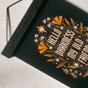 May include: Black wall hanging with the text "HELLO DARKNESS MY OLD FRIEND" in a decorative font. The design includes orange and gold floral elements and a wooden hanging bar.