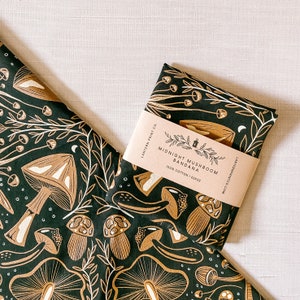 Op de afbeelding: Een zwarte bandana met een wit paddenstoelontwerp en de tekst "Midnight Mushroom Bandana 100% Cotton | 22x22" erop gedrukt.