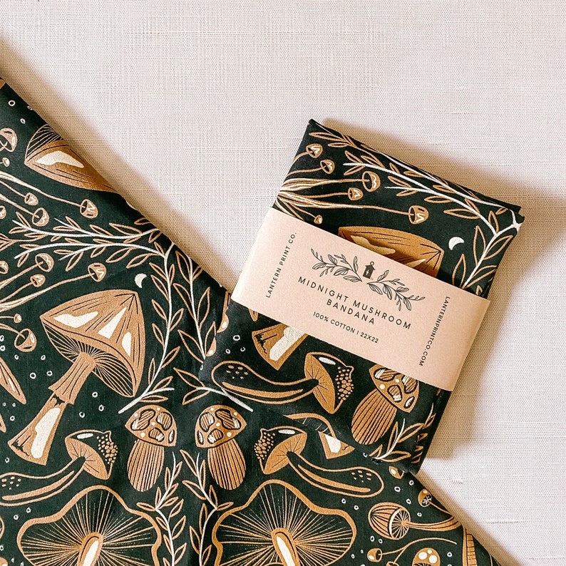 Op de afbeelding: Een zwarte bandana met een wit paddenstoelontwerp en de tekst "Midnight Mushroom Bandana 100% Cotton | 22x22" erop gedrukt.
