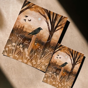 May include: Two art prints featuring a black crow perched on a wooden post in a field of white flowers. The artwork has a warm color palette of browns and tans, with a full moon and flying birds in the background. The prints are of different sizes.