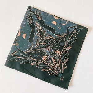 May include: A dark green bandana with a white and gold design featuring a butterfly, mushrooms, stars, and hands. The design is reminiscent of a mystical forest scene.