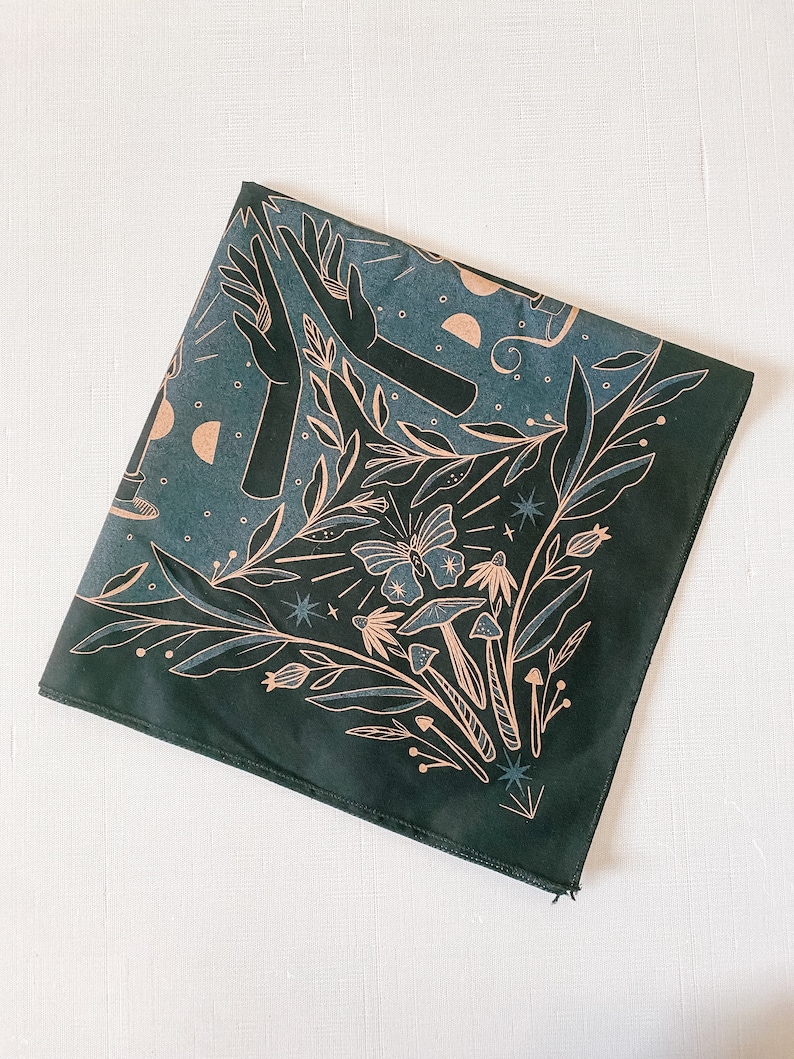 May include: A dark green bandana with a white and gold design featuring a butterfly, mushrooms, stars, and hands. The design is reminiscent of a mystical forest scene.