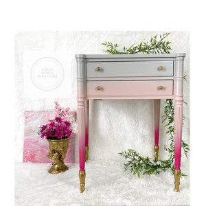 May include: A two-drawer side table with a gray to pink ombre paint finish. The table has gold-colored legs and hardware. The table is sitting on a white fluffy surface.