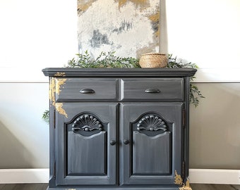 May include: A gray painted wooden cabinet with gold accents. The cabinet has two drawers and two doors with carved details. The cabinet is sitting on a wood floor.