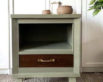 May include: A sage green nightstand with a single drawer and a shelf. The drawer has a dark wood front and a gold metal handle. The nightstand has four legs and is sitting on a patterned rug.