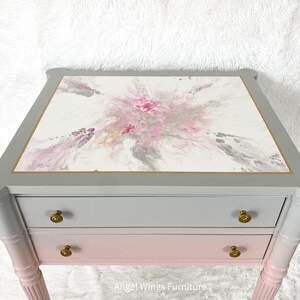 May include: A two-drawer end table with a gray and pink ombre finish. The top drawer is gray and the bottom drawer is pink. The top of the table has a white surface with a pink and gray abstract design. The table has gold-colored hardware and turned legs.