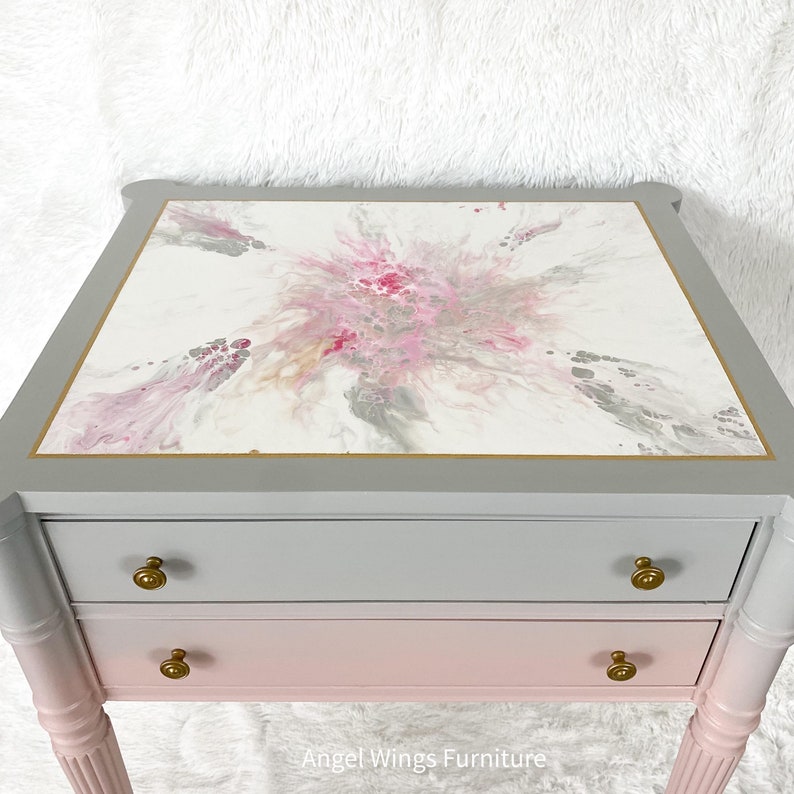 May include: A two-drawer end table with a gray and pink ombre finish. The top drawer is gray and the bottom drawer is pink. The top of the table has a white surface with a pink and gray abstract design. The table has gold-colored hardware and turned legs.