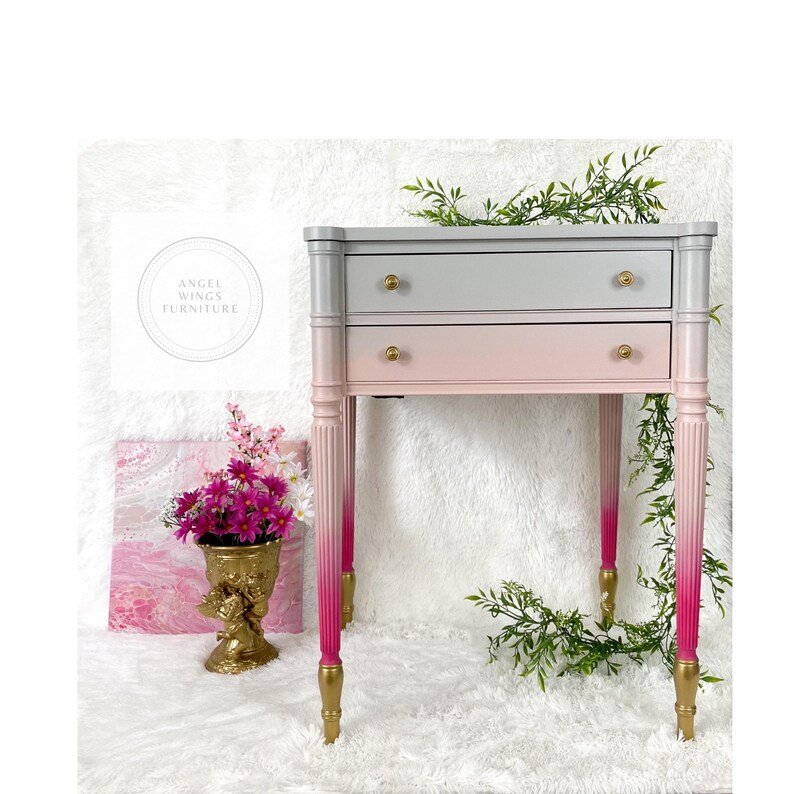 May include: A two-drawer side table with a gray to pink ombre paint finish. The table has gold-colored legs and hardware. The table is sitting on a white fluffy surface.