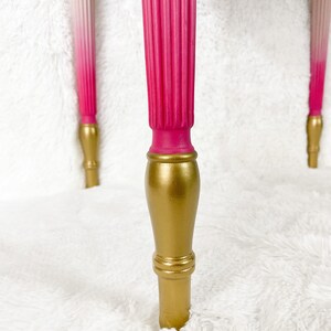 May include: Close-up of a table leg with a pink to white ombre paint finish and a gold base. The leg is on a white fluffy surface.  Angel Wings Furniture.