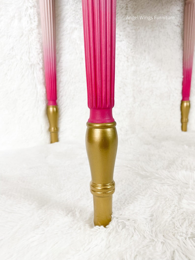 May include: Close-up of a table leg with a pink to white ombre paint finish and a gold base. The leg is on a white fluffy surface.  Angel Wings Furniture.
