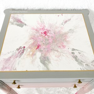 May include: A gray painted end table with two drawers and gold trim. The top of the table is painted with a pink, gray, and white abstract design. The text "Angel Wings Furniture" is visible on the top of the table.