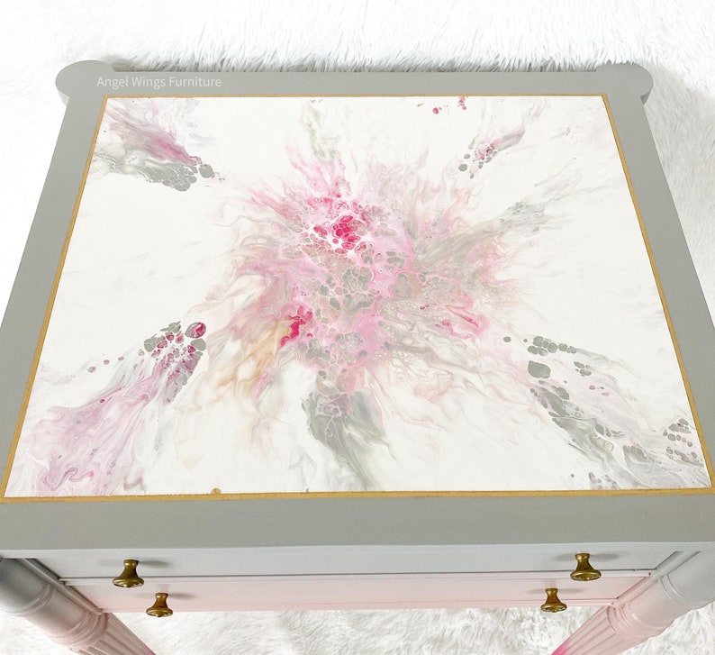 May include: A gray painted end table with two drawers and gold trim. The top of the table is painted with a pink, gray, and white abstract design. The text "Angel Wings Furniture" is visible on the top of the table.