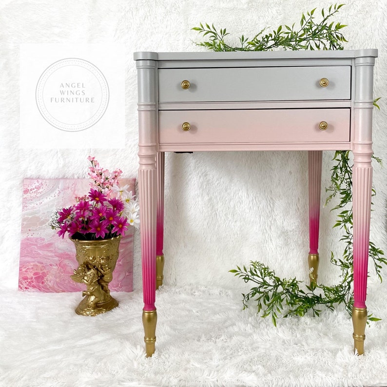 May include: A two-drawer side table with a gray to pink ombre paint finish. The table has gold-painted legs with a pink ombre effect. The table is on a white fluffy surface.