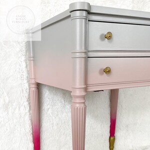 May include: A two-drawer side table with a ombre finish, transitioning from silver to pink. The table has turned legs with a fluted design and gold accents on the drawer pulls.