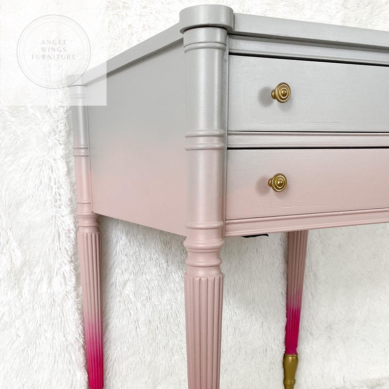 May include: A two-drawer side table with a ombre finish, transitioning from silver to pink. The table has turned legs with a fluted design and gold accents on the drawer pulls.