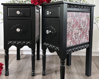 May include: Two black nightstands with ornate details and silver drawer pulls. The nightstand on the right has a floral and skull design on the front.