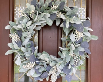 May include: A circular wreath made of faux greenery, featuring large, soft, gray-green leaves and accents of small white flowers. The wreath is hanging on a brown door with glass panels.