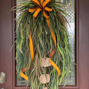 May include: A fall-themed wreath with green and brown faux foliage, an orange bow, and three wooden pumpkin ornaments. The wreath is hanging on a brown door with a glass window.