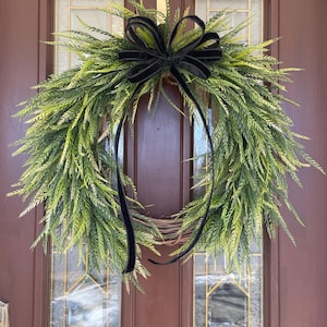 May include: A green wreath with a black velvet bow. The wreath is made of artificial greenery and is hanging on a brown door.