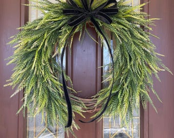 May include: A green wreath with a black velvet bow. The wreath is made of artificial greenery and is hanging on a brown door.