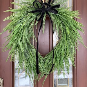 May include: A green fern wreath with a black velvet ribbon bow. The wreath is hanging on a brown door.