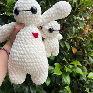 May include: Two white crochet plush toys, one large and one small, resembling a character with black oval eyes and a red heart. The larger toy has outstretched arms and a rounded body. The smaller toy is held next to the larger one.