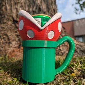 Può includere: Una tazza in 3D stampata in verde e rosso a forma di pianta Piranha di Super Mario Bros. La tazza ha un bordo bianco e punti bianchi. Una lattina verde è all'interno della tazza.