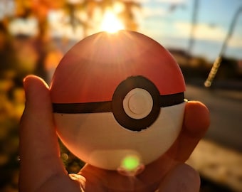 Op de afbeelding: Een rood en wit Pokeball-speelgoed met een zwarte ring in het midden. Het speelgoed wordt in een hand gehouden met een wazige achtergrond van herfstbladeren en een zonsondergang.