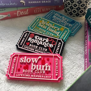 May include: A collection of books, including "The Fine Print" and "Icebreaker", sits beside a small potted plant. In front are ticket-shaped magnets, with text such as "Dark Romance Book Club" and "Slow Burn Book Club".