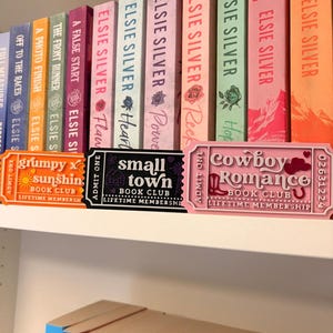 May include: Three rectangular plastic ticket-shaped signs labeled Book Club Lifetime Membership. The orange sign reads Grumpy x Sunshine, the black sign reads Small Town, and the pink sign reads Cowboy Romance. They are displayed in front of a row of colorful books.