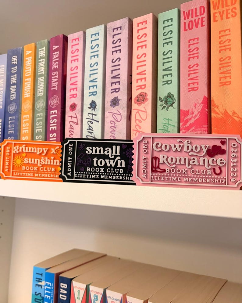 May include: Three rectangular plastic ticket-shaped signs labeled Book Club Lifetime Membership. The orange sign reads Grumpy x Sunshine, the black sign reads Small Town, and the pink sign reads Cowboy Romance. They are displayed in front of a row of colorful books.
