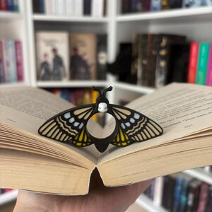 May include: A black and yellow moth-shaped book page holder sits on an open book. The book page holder has a white skull design on the top. The wings have yellow and white accents. The background is a bookshelf filled with books.
