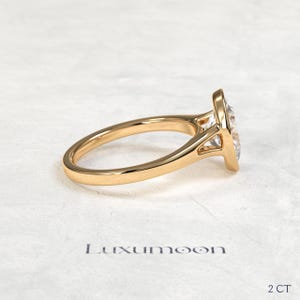 May include: A gold ring featuring a clear, pear-shaped stone set in a polished metal band. The side profile shows the setting structure. The text Luxumoon and 2 CT are printed below the ring, indicating the brand name and a 2 carat weight.