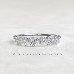 May include: A silver-toned ring featuring a cluster of multiple round, clear, faceted stones arranged in a horizontal row across the top. The word Luxumoon is printed in dark blue letters below the ring on the textured white surface.