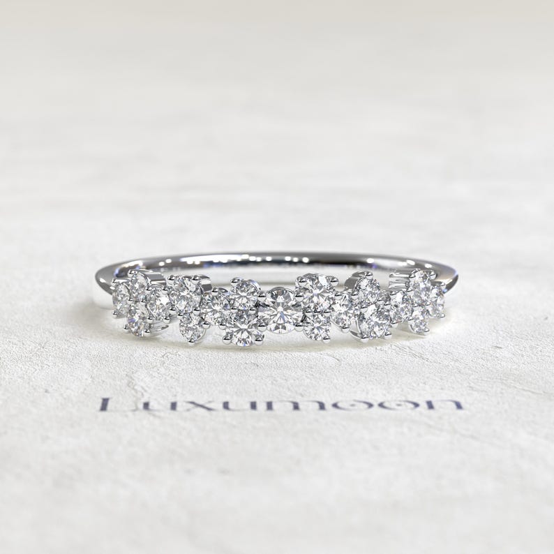May include: A silver-toned ring featuring a cluster of multiple round, clear, faceted stones arranged in a horizontal row across the top. The word Luxumoon is printed in dark blue letters below the ring on the textured white surface.