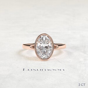 May include: A rose gold ring featuring a large oval-cut clear gemstone set in a bezel setting. The band has a split shank design near the center stone. The text Luxumoon is printed below the ring, and the text 2 CT is printed in the bottom right.