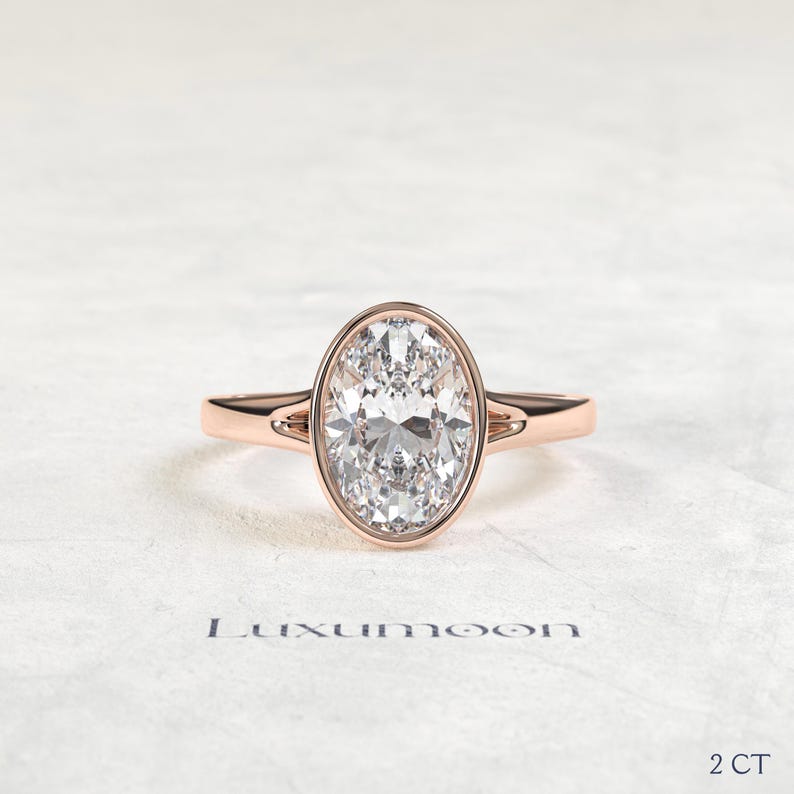 May include: A rose gold ring featuring a large oval-cut clear gemstone set in a bezel setting. The band has a split shank design near the center stone. The text Luxumoon is printed below the ring, and the text 2 CT is printed in the bottom right.