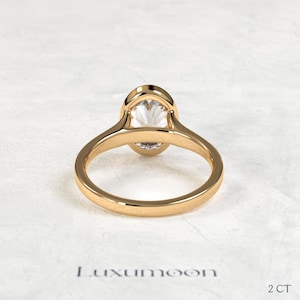 May include: A gold ring featuring a single oval-cut clear gemstone set in a smooth bezel setting. The band is polished gold with a rounded profile. The text Luxumoon and 2 CT are visible at the bottom of the frame, indicating the brand and weight.