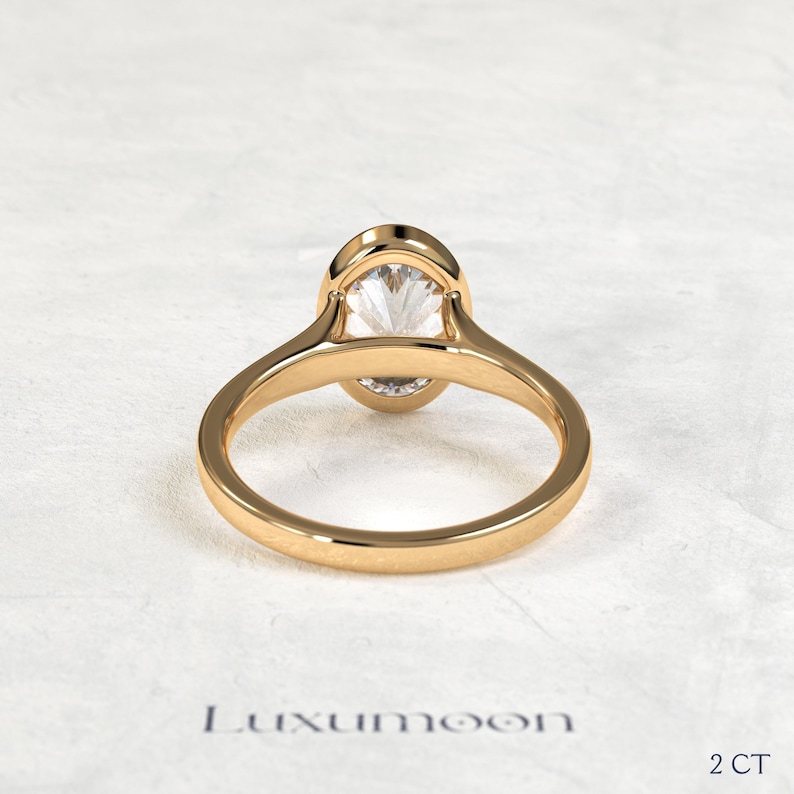 May include: A gold ring featuring a single oval-cut clear gemstone set in a smooth bezel setting. The band is polished gold with a rounded profile. The text Luxumoon and 2 CT are visible at the bottom of the frame, indicating the brand and weight.
