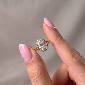 May include: A gold ring featuring a large, clear, oval-cut gemstone set in a smooth, polished bezel setting. The band is thin and circular, crafted from a warm-toned yellow metal that complements the faceted, brilliant-cut center stone.
